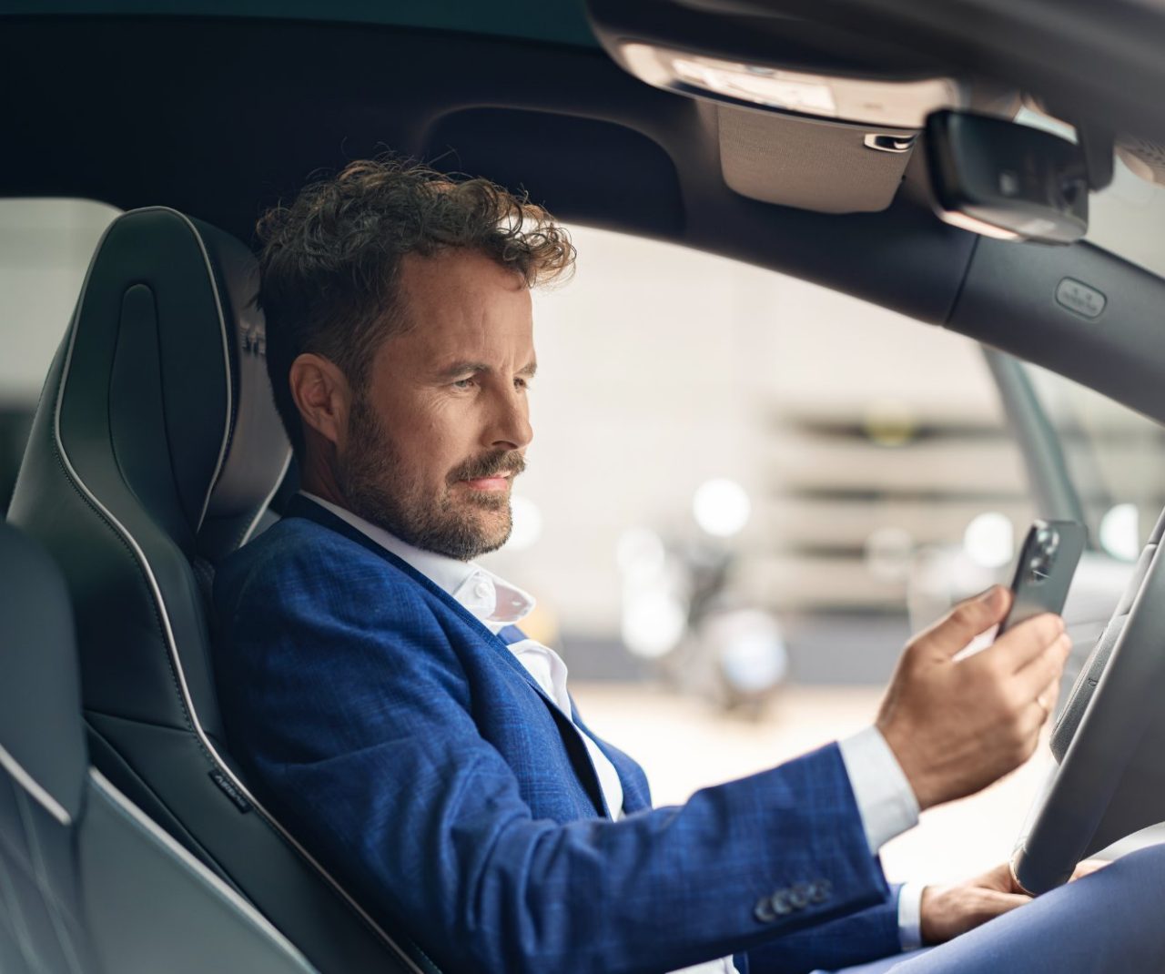Homme assis au volant d'une voiture, regardant son téléphone, vu depuis la porte conducteur.