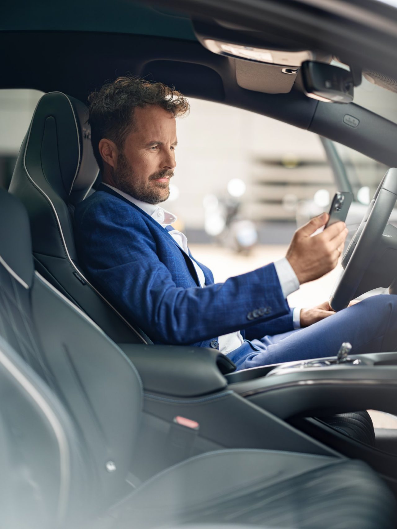 Homme assis au volant d'une voiture, regardant son téléphone, vu depuis la porte conducteur.