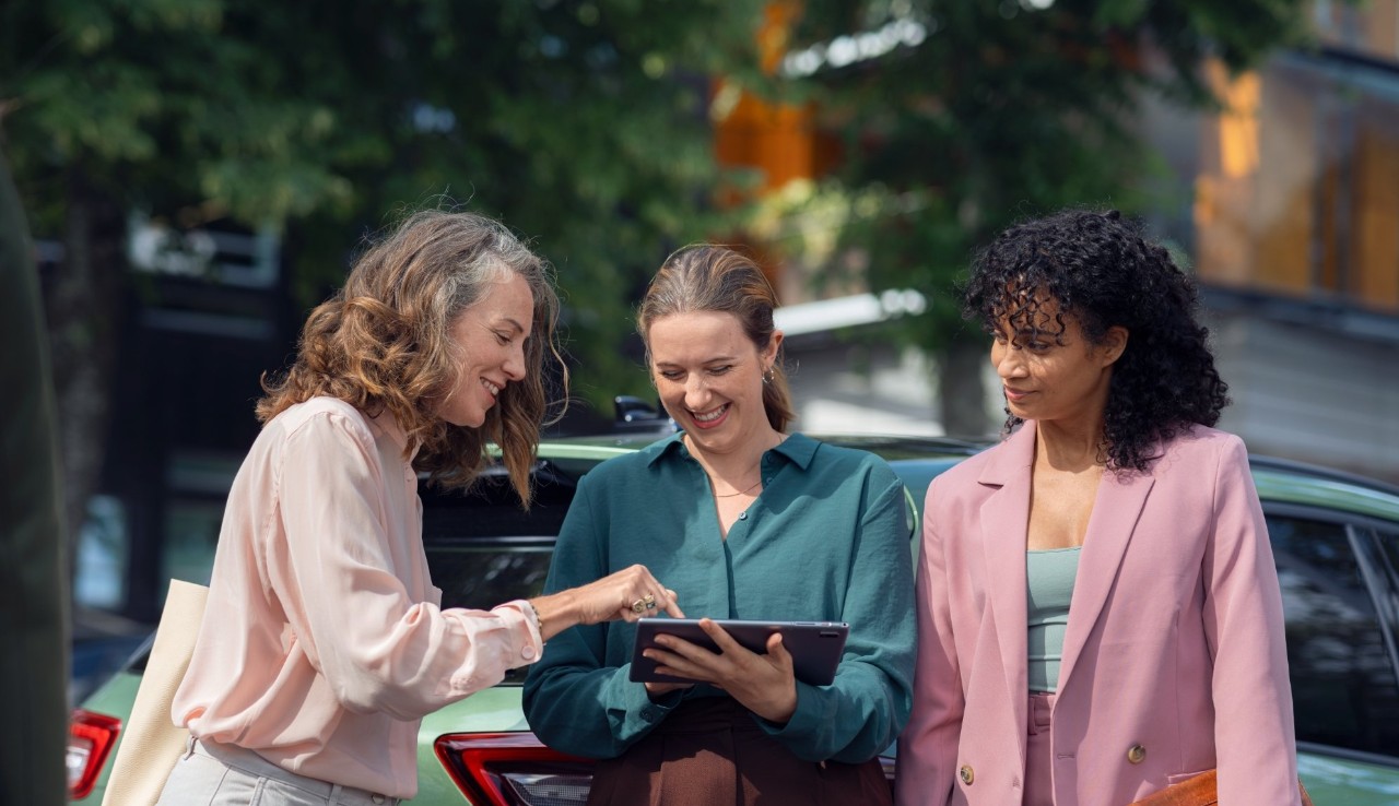 Trois femmes discutent devant une voiture verte, tenant une tablette.