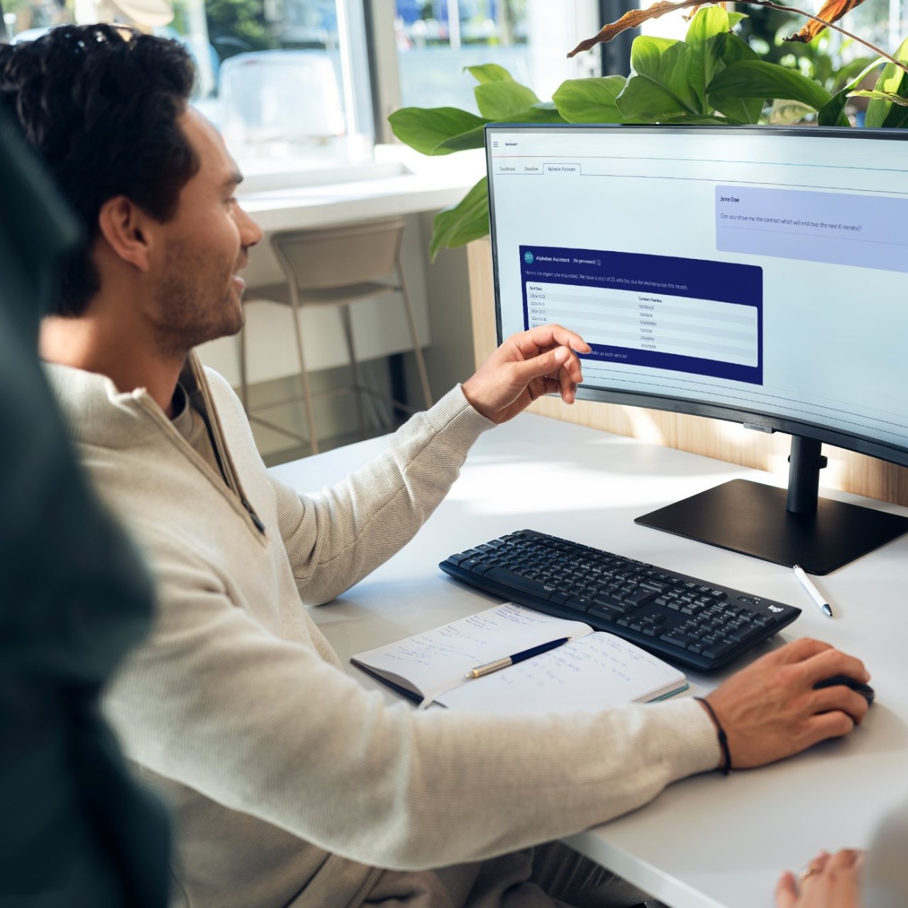 Personne assise à un bureau dans un bureau moderne, interagissant avec des données sur un écran large, avec un ordinateur portable et un clavier sur la table et des plantes vertes en arrière-plan.