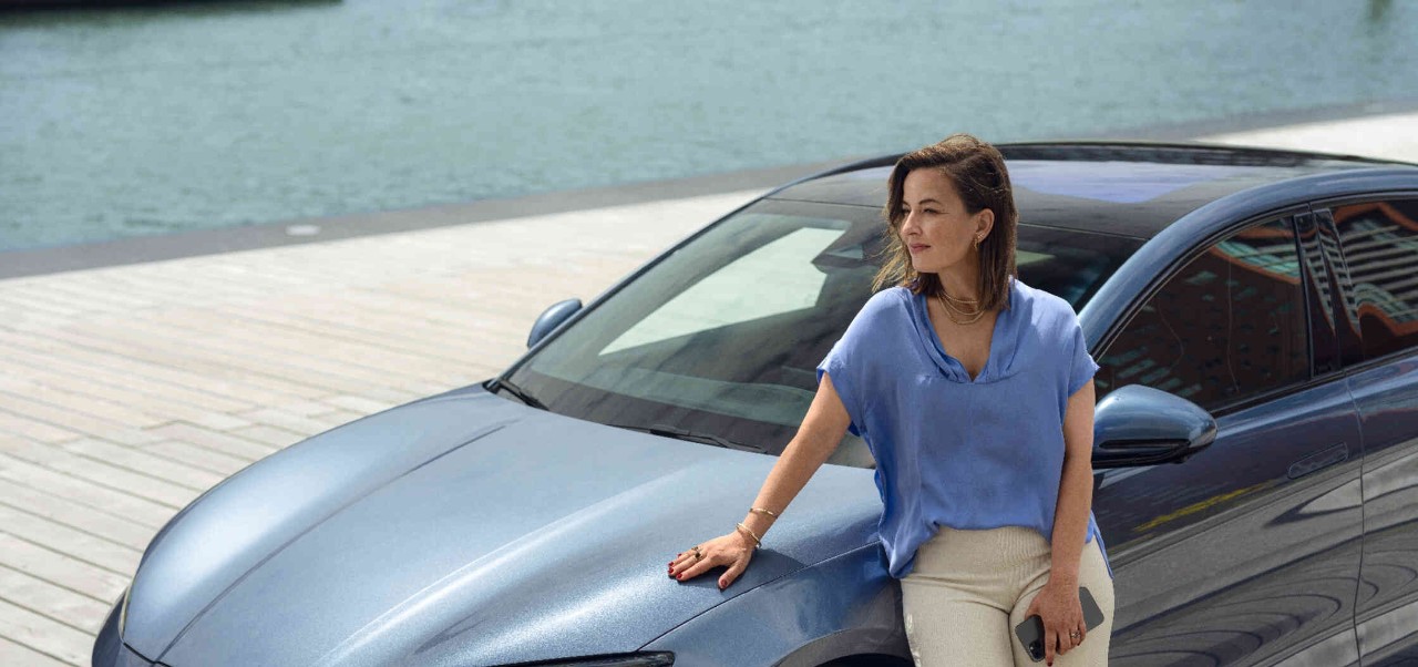 Eine Frau lehnt sich an ein blaugraues BYD Auto und schaut in die Ferne w?hrend im Hintergrund ein Fluss und st?dtische Architektur sichtbar sind.