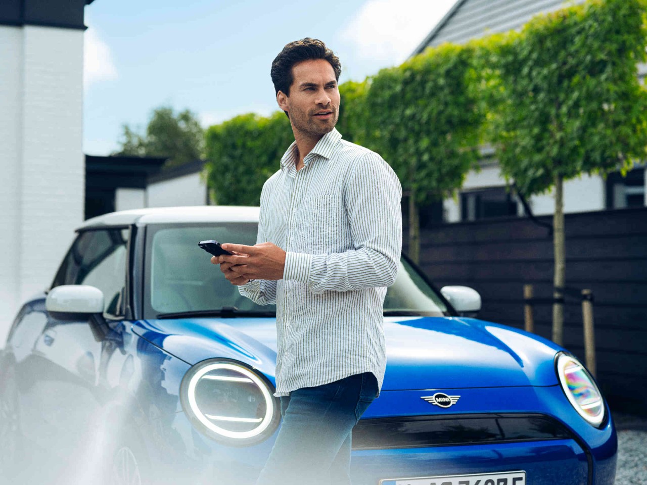 Man houdt zijn telefoon in de hand kijkt naar links en staat voor een blauwe MINI-auto.