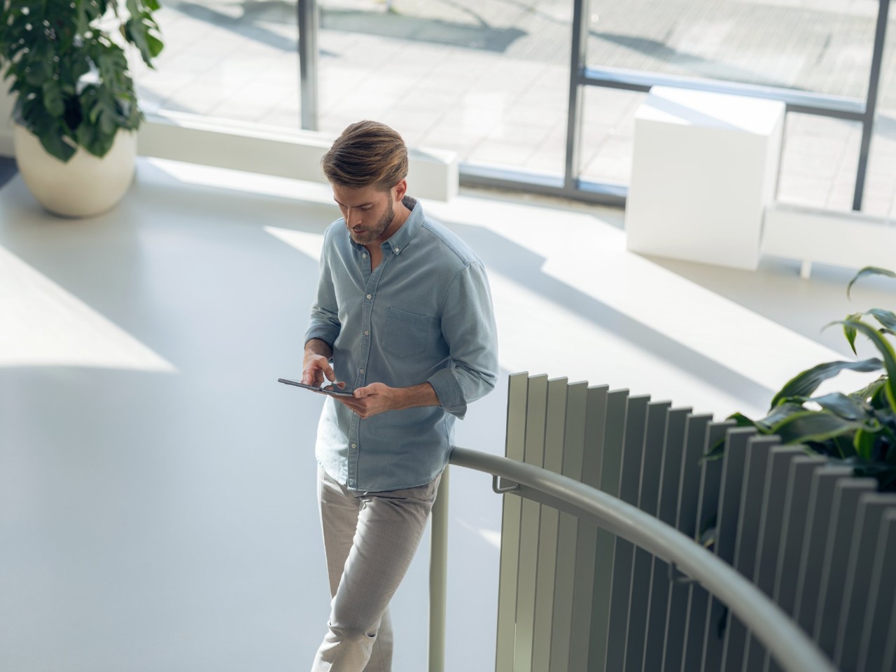 Een man leunt tegen de leuning van een trap met een tablet in een moderne, goed verlichte ruimte.