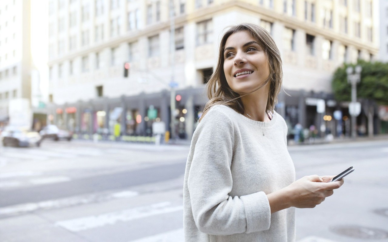 Vrouw met een witte trui bij een groot kruispunt met telefoon in haar hand.