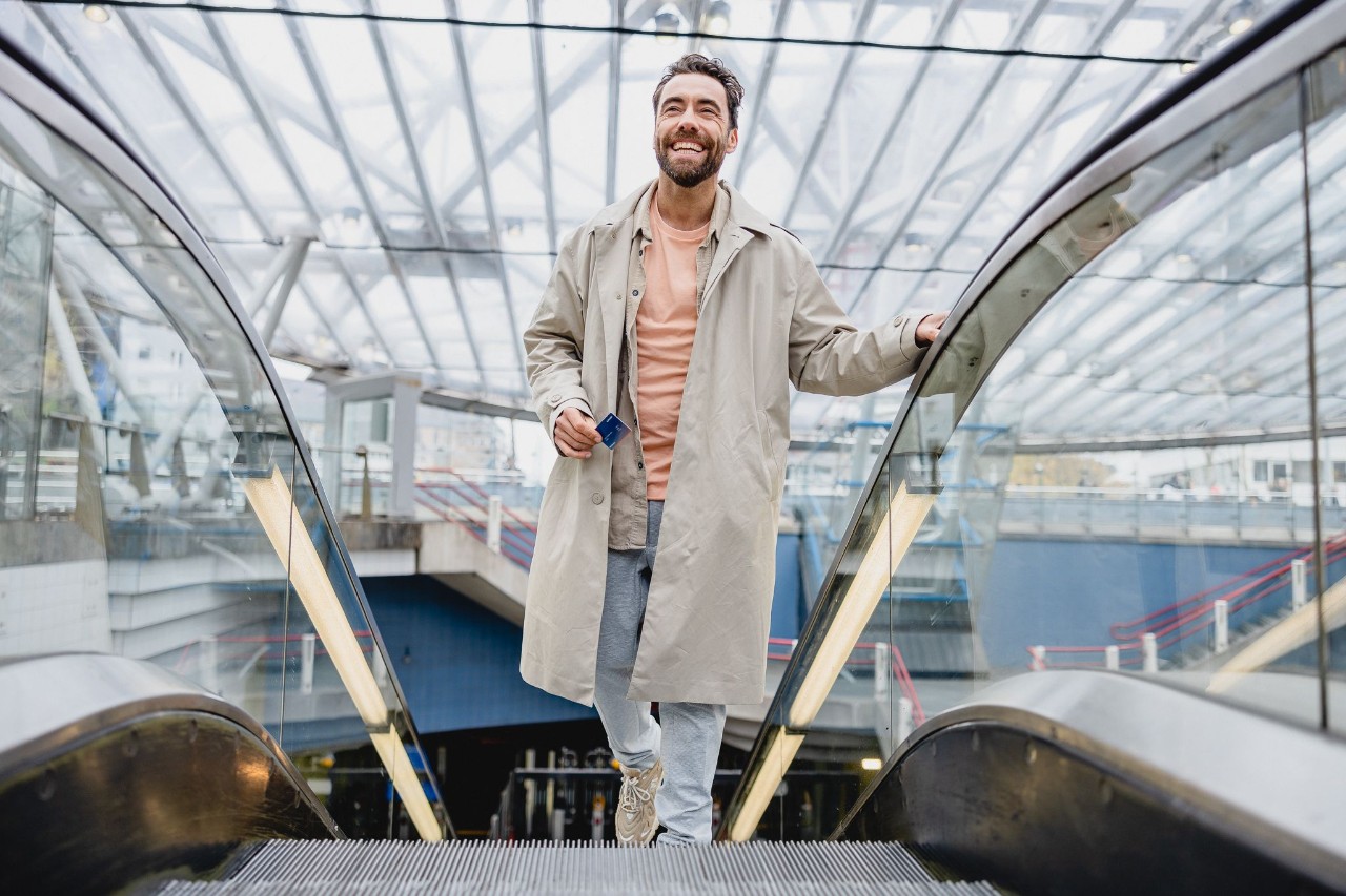 Man in een lange grijze jas staat op een roltrap.