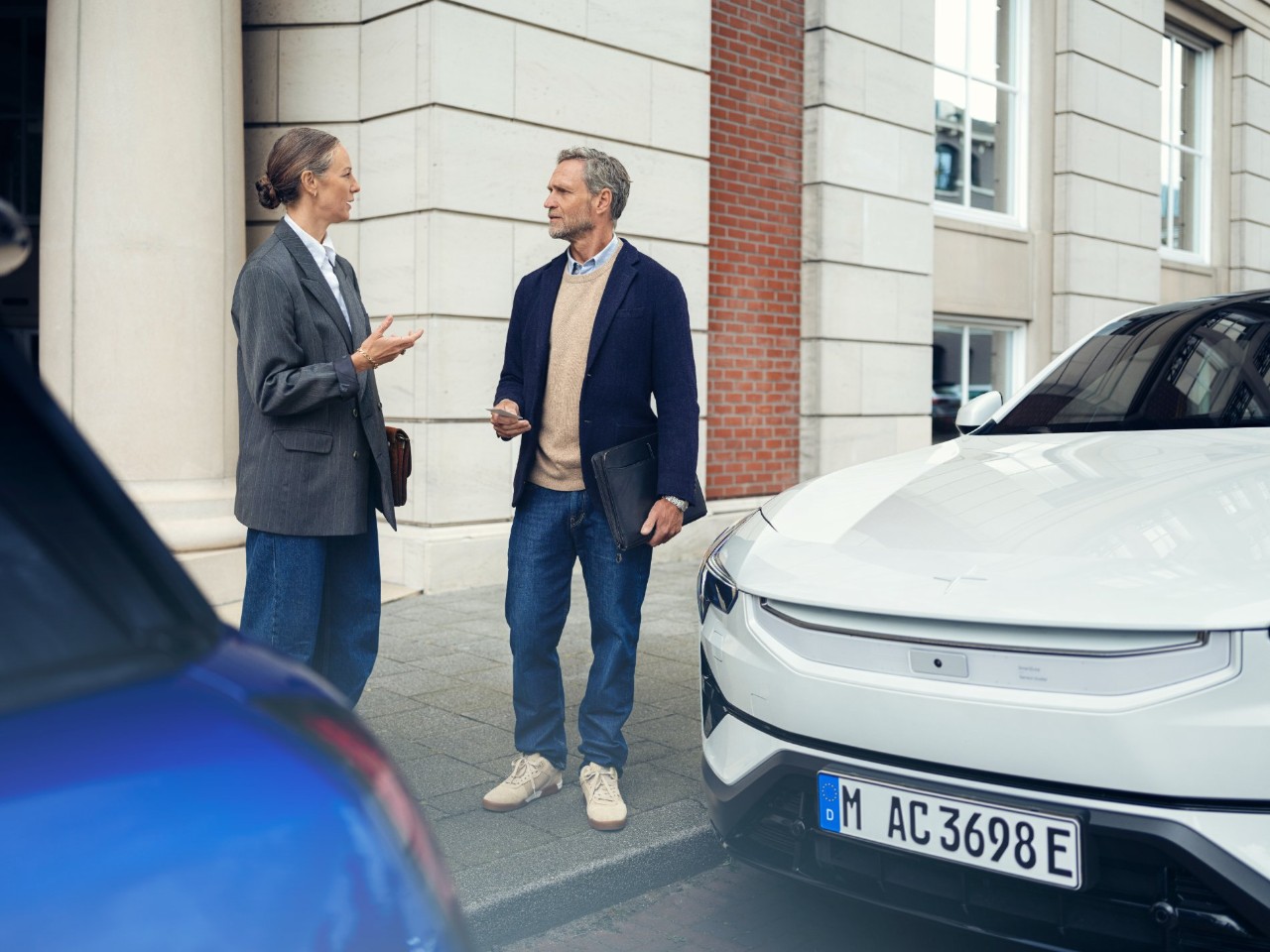 Een vrouw en een man voeren een gesprek op de stoep voor een elegant gebouw, naast twee geparkeerde auto's.