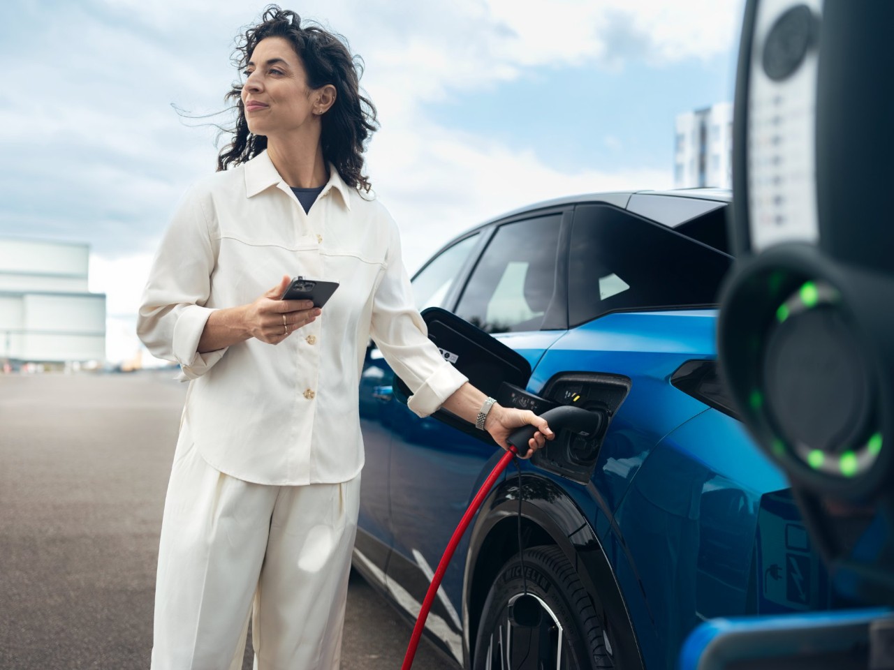 Een vrouw houdt een oplaadkabel en een smartphone vast naast een blauwe elektrische auto bij een laadstation.