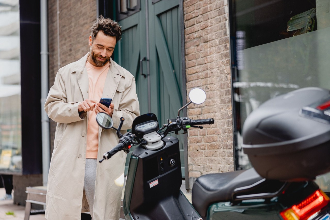 Man voor een deur bij een scooter kijkend naar zijn telefoon