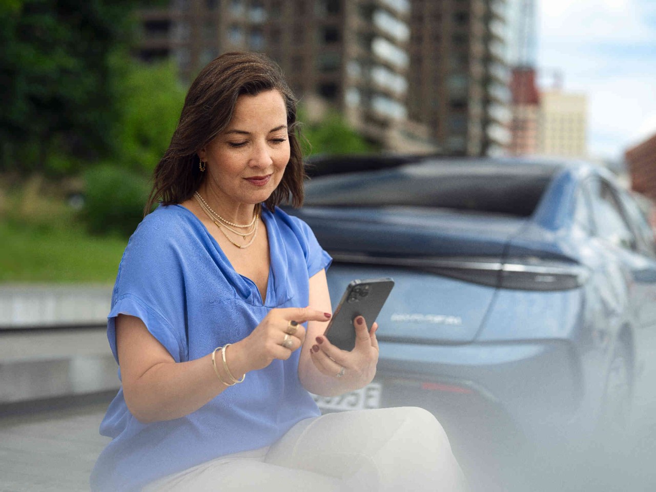 Vrouw zit en kijkt op haar telefoon achter een blauw auto.