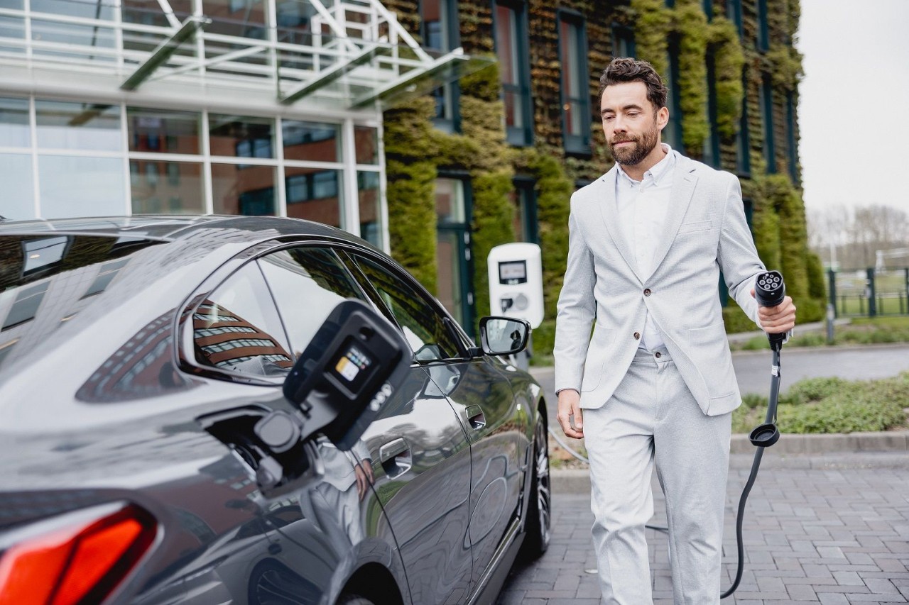 Een man in een lichtkleurig pak staat naast een elektrische auto en houdt een laadkabel vast bij een laadpaal voor een modern gebouw.
