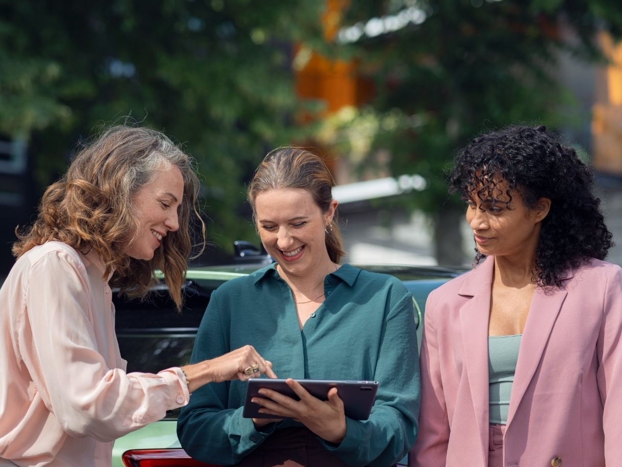 Drie vrouwen overleggen voor een groene auto, terwijl ze een tablet vasthouden.