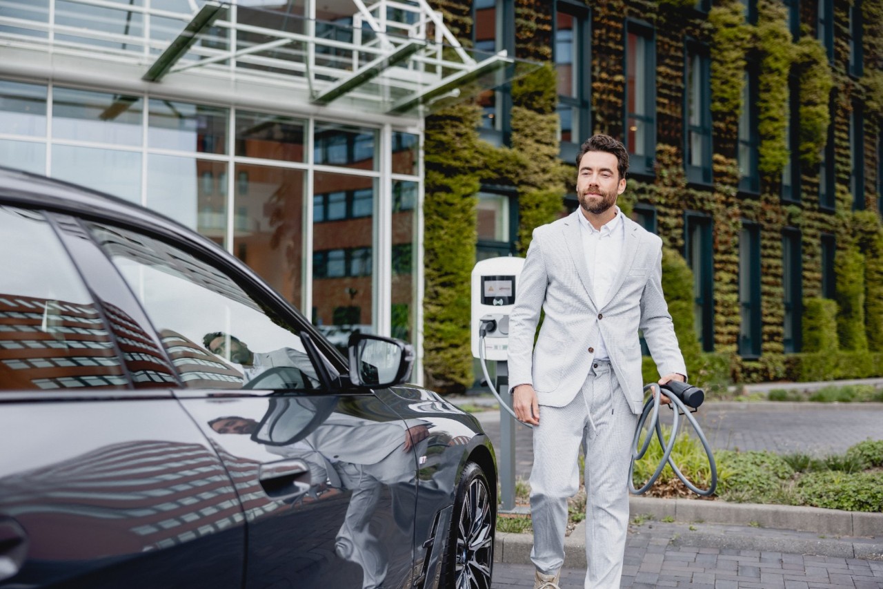 Een man in een lichtkleurig pak staat naast een elektrische auto en houdt een laadkabel vast bij een laadpaal voor een modern gebouw.