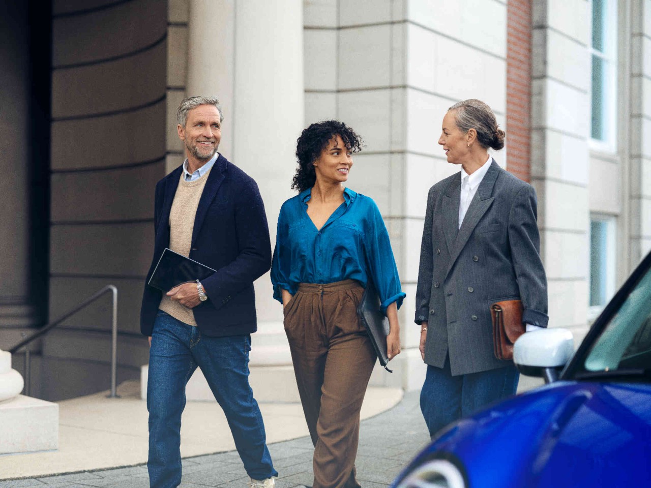 Drie personen lopen op een stoep voor een elegant gebouw man draagt een tablet vrouwen dragen aktetassen.