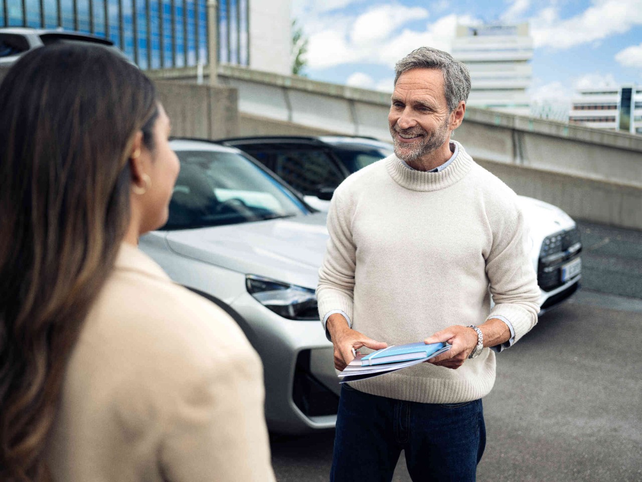 Man toont documenten aan een vrouw op parkeerplaats zilveren en witte auto op de achtergrond.