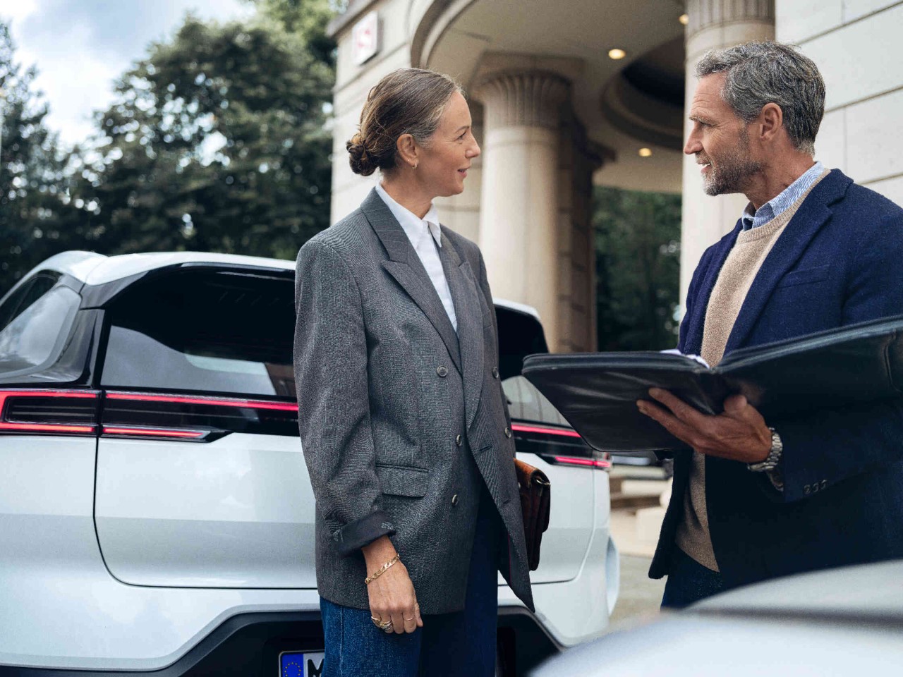 Vrouw en man converseren tussen twee geparkeerde auto's voor een elegant gebouw met zuilen.