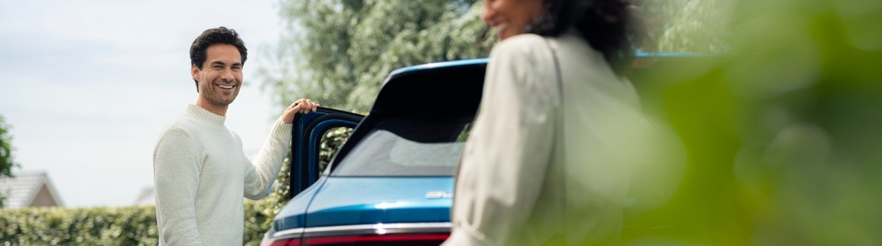 Een man glimlacht naast een blauwe auto en houdt de portier open