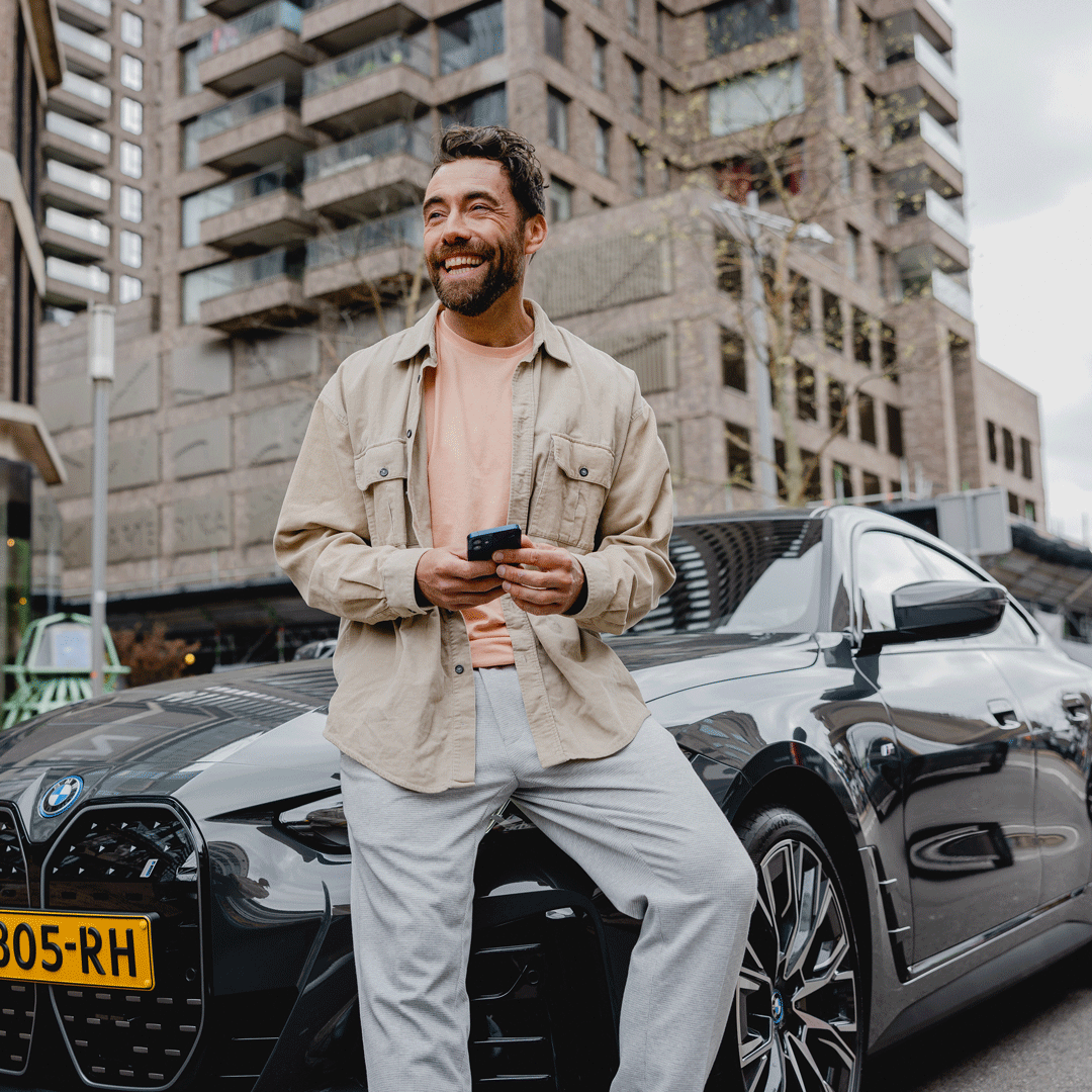 Lachende man zittend op motorkap van BMW met telefoon in zijn hand