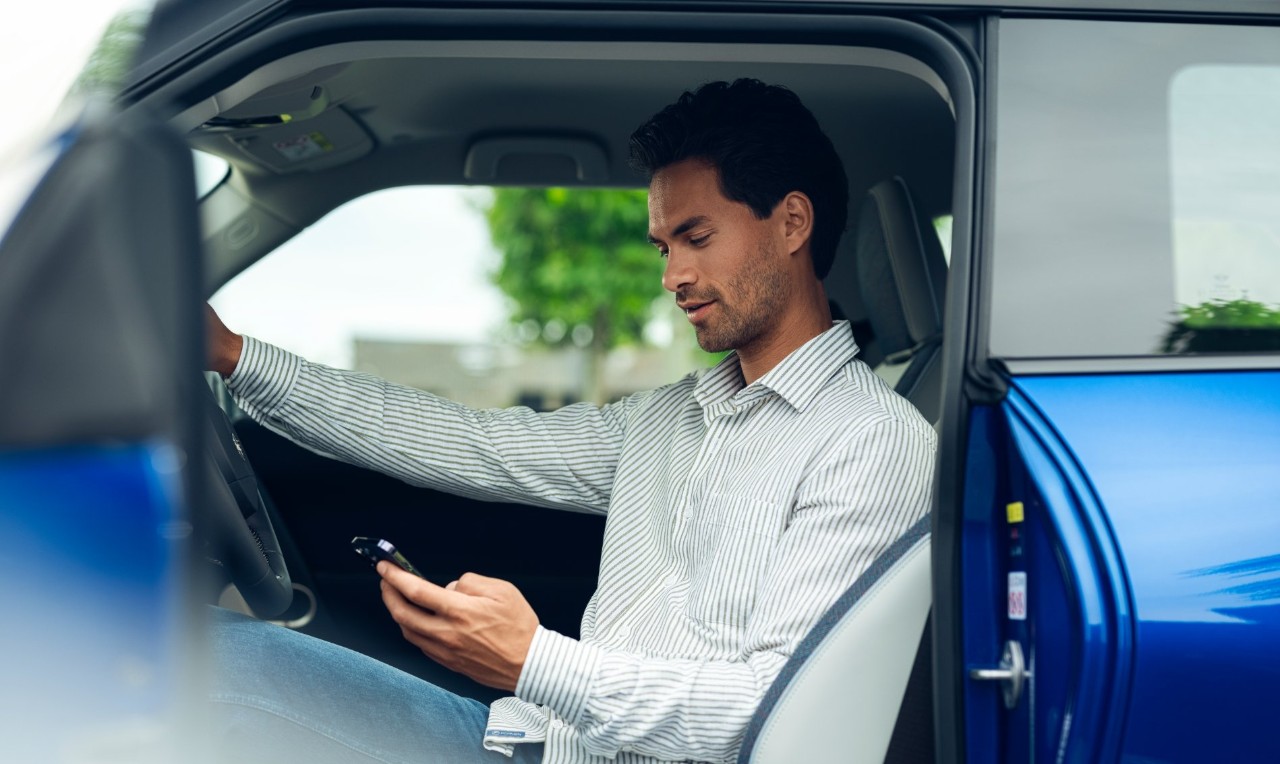 Een man zit op de bestuurdersstoel van een stilstaande auto en kijkt op zijn telefoon, met het bestuurdersportier open.