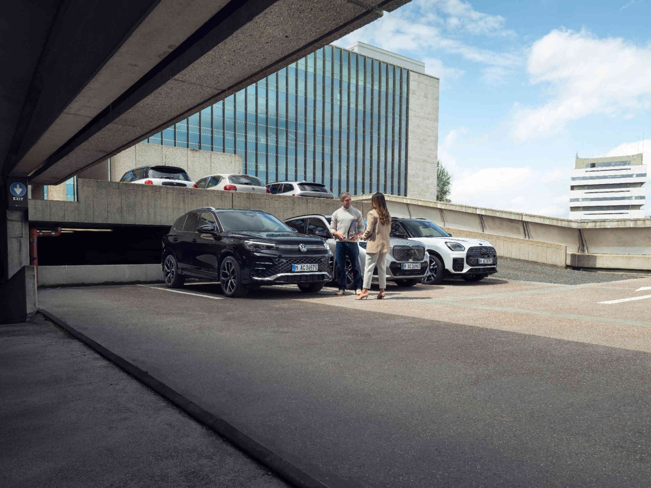 Twee personen converseren tussen geparkeerde auto's in een parkeergarage voor een modern kantoorgebouw.