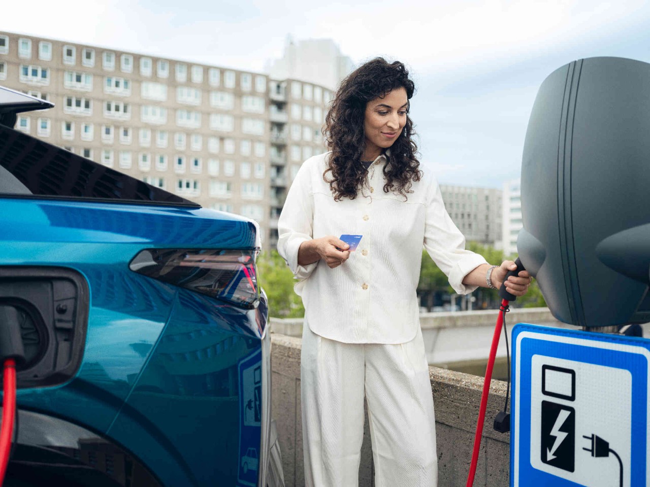 Vrouw laadt elektrische voertuig aan laadstation houdt blauwe kaart vast rood laadkabel zichtbaar.