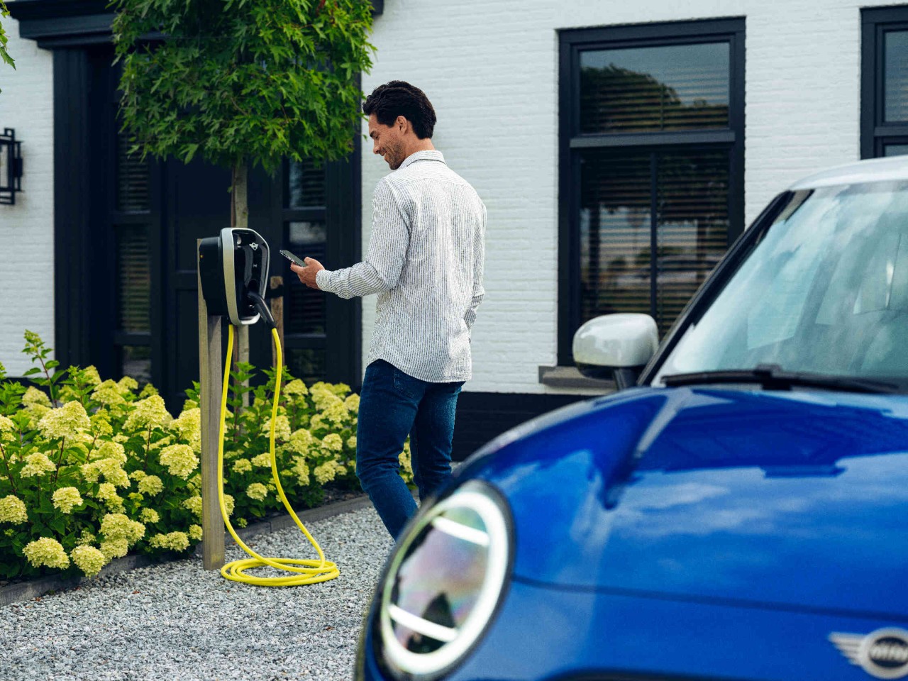 Man kijkt op zijn telefoon en staat voor een laadstation achter hem een blauwe MINI.