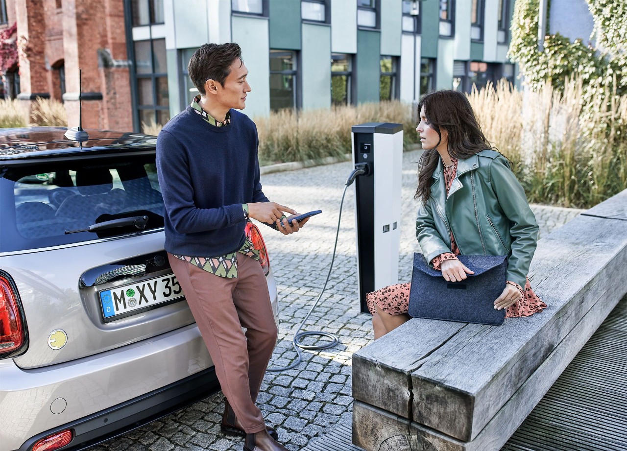 Een vrouw houdt een oplaadkabel en een smartphone vast naast een blauwe elektrische auto bij een laadstation.