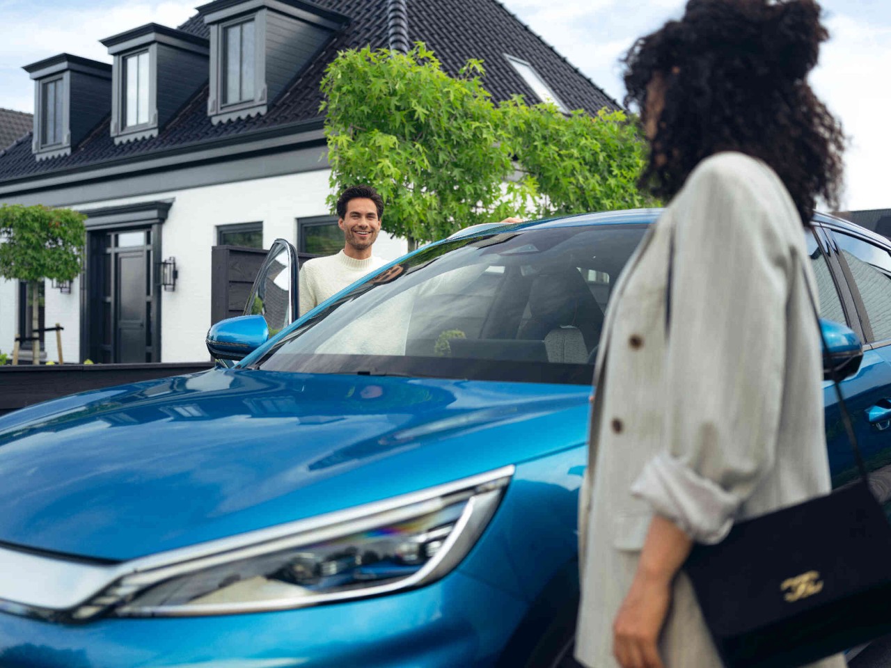 Blauwe auto voor een elegant huis vrouw in lichte kleding met tas op de voorgrond.