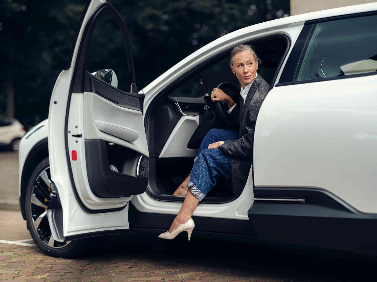 Vrouw zit op de bestuurdersstoel van een geparkeerde witte auto en kijkt met een open deur naar links over haar schouder.