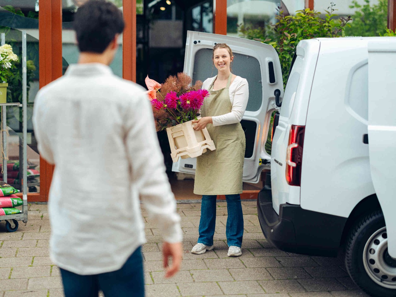 Floriste met een mand vol kleurrijke bloemen voor de bloemenwinkel man loopt naar haar toe.