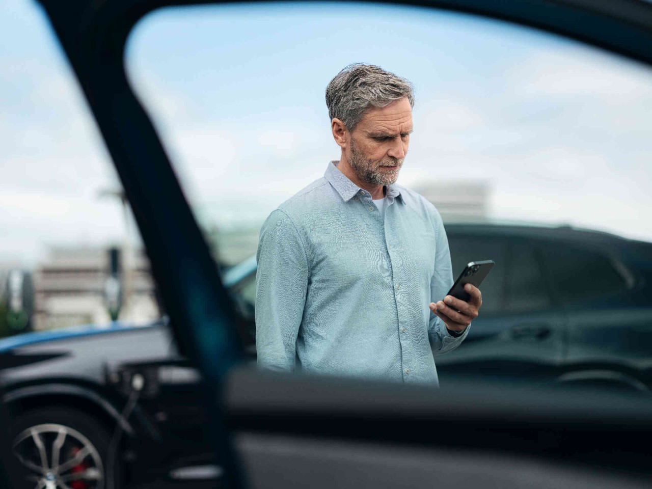 Man in een lichtblauw shirt houdt een smartphone vast staat naast een zwarte auto op een parkeerplaats.