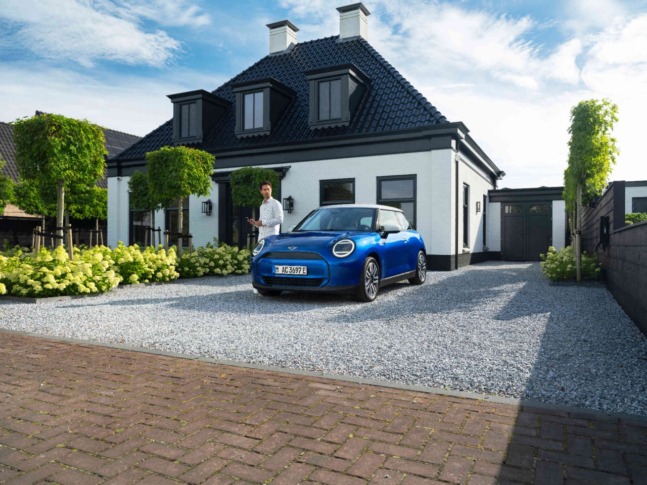 Man staat links naast een blauwe MINI-auto op een grindoprit voor een woonhuis.