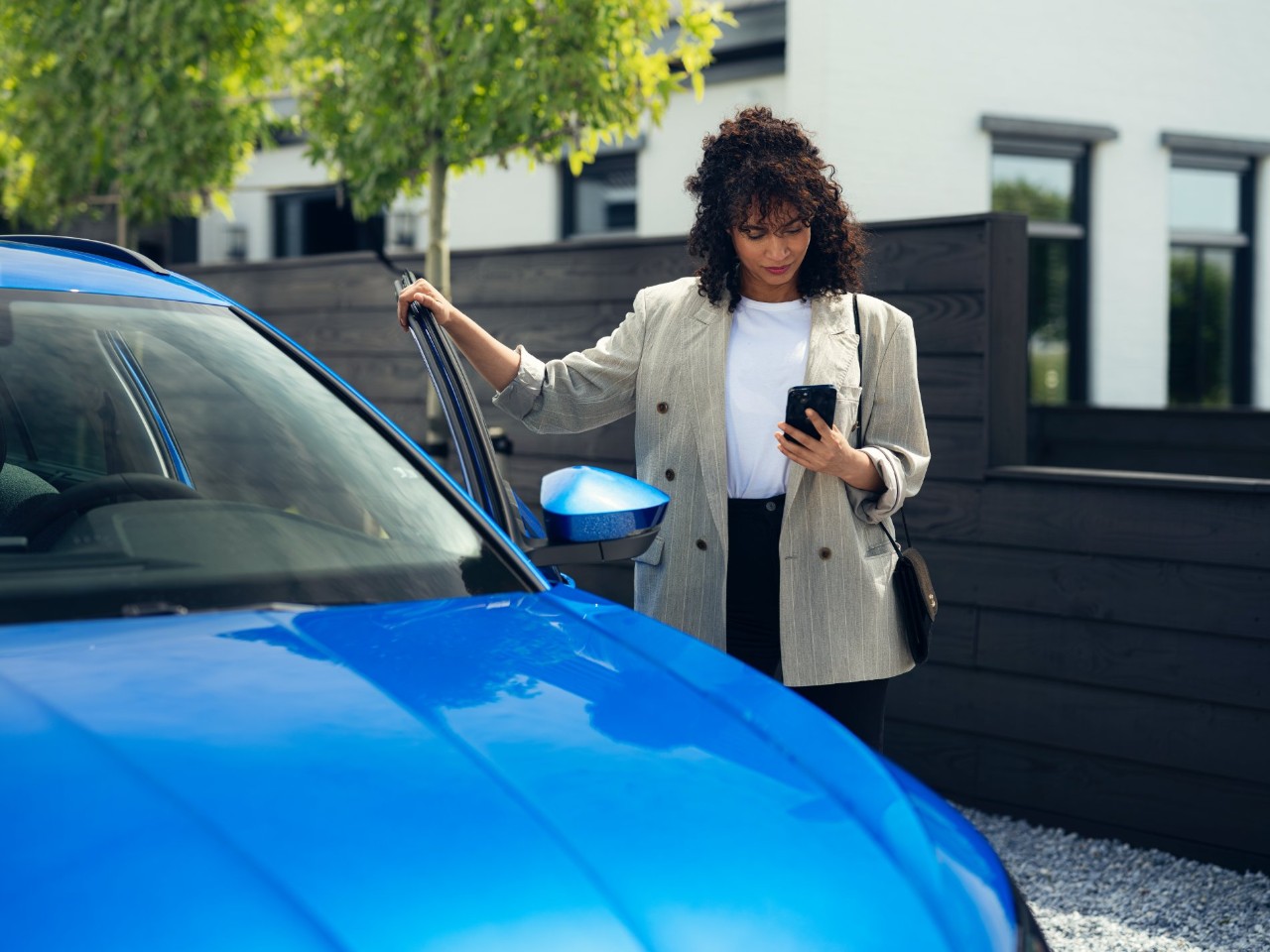 Vrouw die naast een blauwe auto staat, een smartphone vasthoudt en het bestuurdersportier opent in een stedelijke woonomgeving.