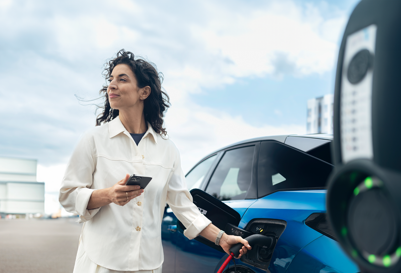 Vrouw die naast een blauwe auto staat, een smartphone vasthoudt en het bestuurdersportier opent in een stedelijke woonomgeving.