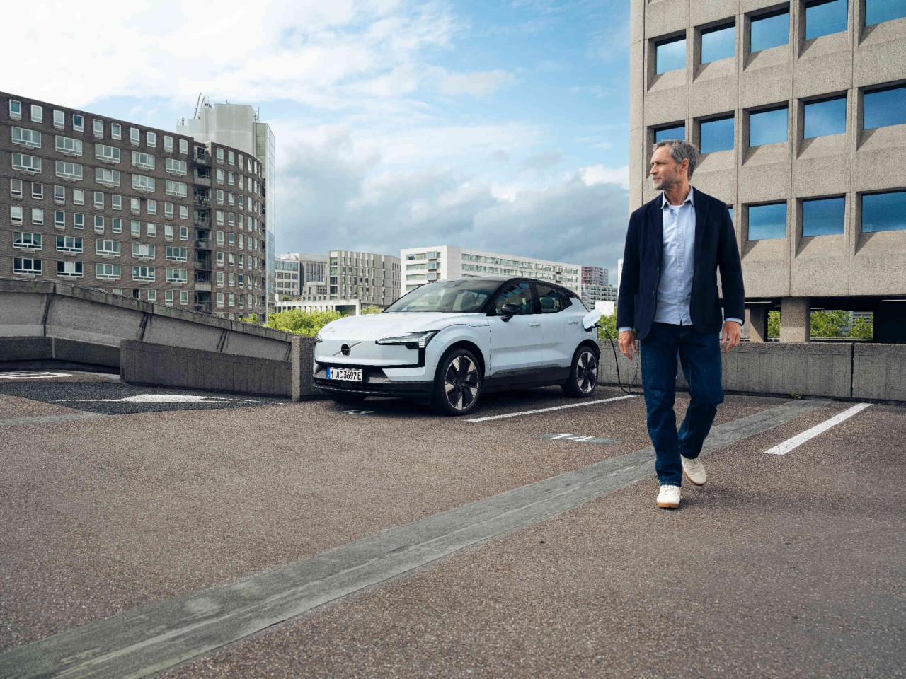 Witte elektrische auto Volvo aan laadstation op stedelijk parkeerterrein moderne gebouwen op de achtergrond.