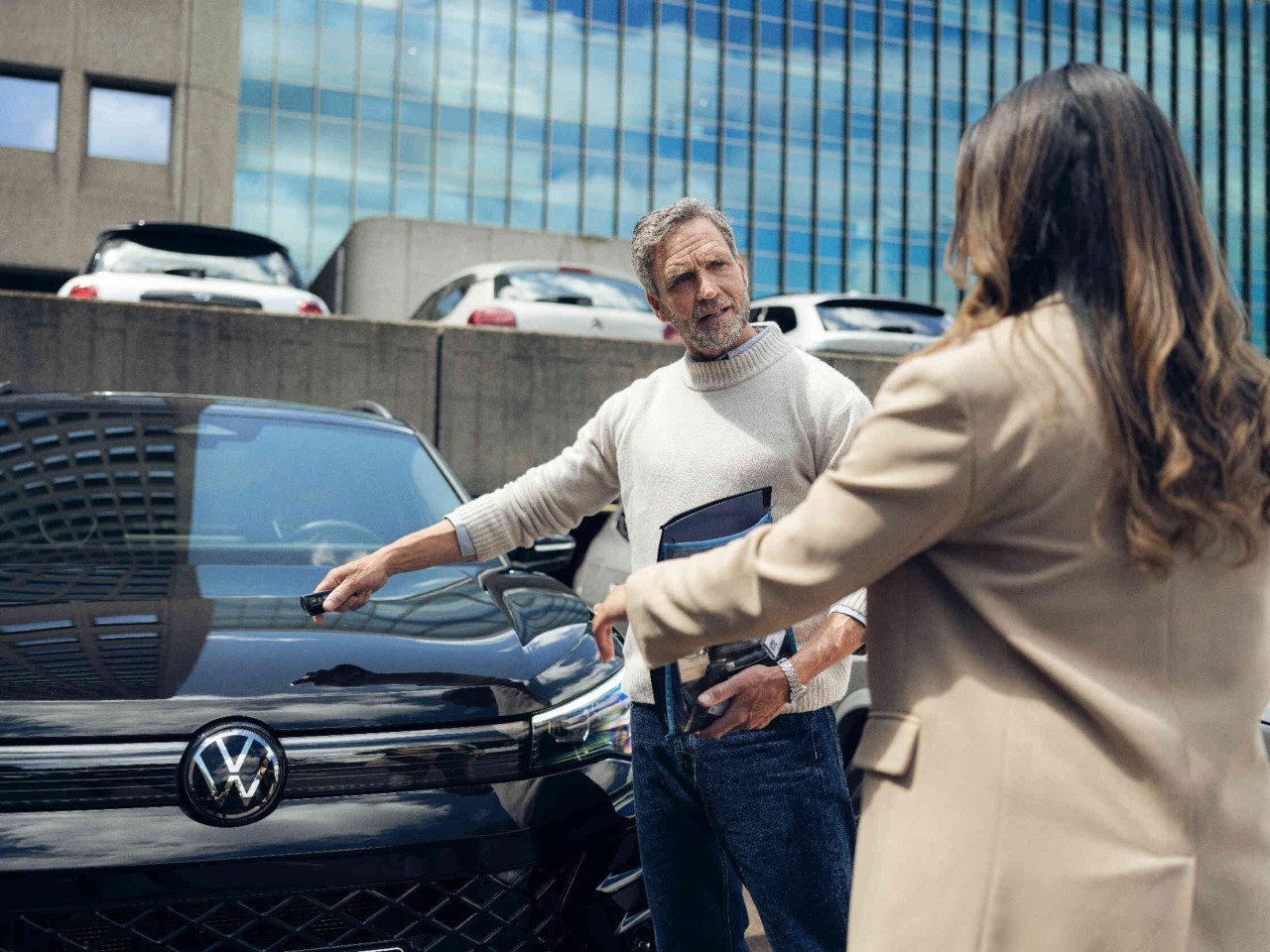 Man wijst naar zwarte Volkswagen terwijl hij met een vrouw praat voor een modern gebouw.