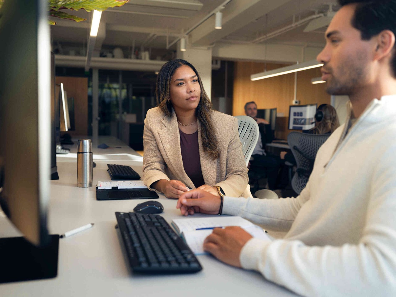 Man aan een bureau met monitor en notitieboek vrouw in blazer interageert moderne kantooromgeving.
