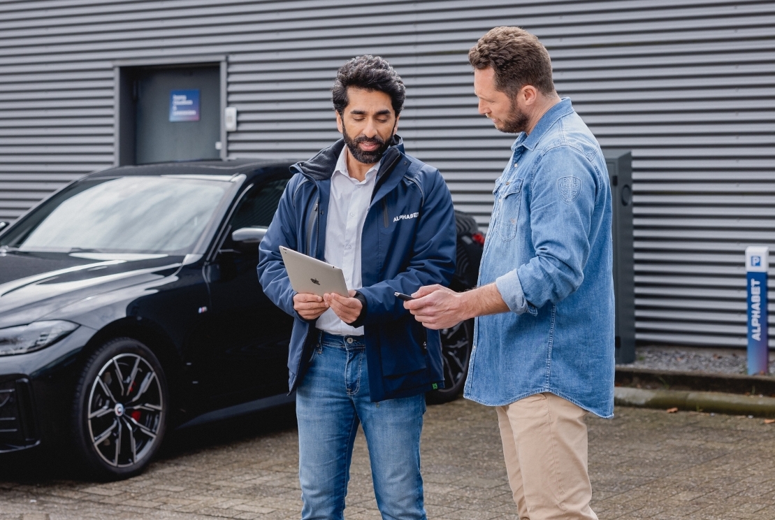 Mann sitzt auf Fahrersitz eines stehendes Autos schaut auf sein Handy Fahrertuer steht offen.